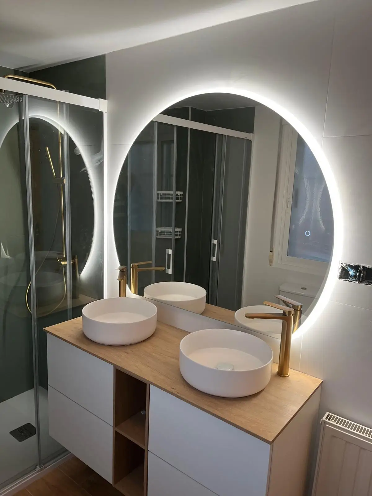 Reforma de baño con mueble de madera, lavabos sobre encimera y espejo circular retroiluminado en Madrid
