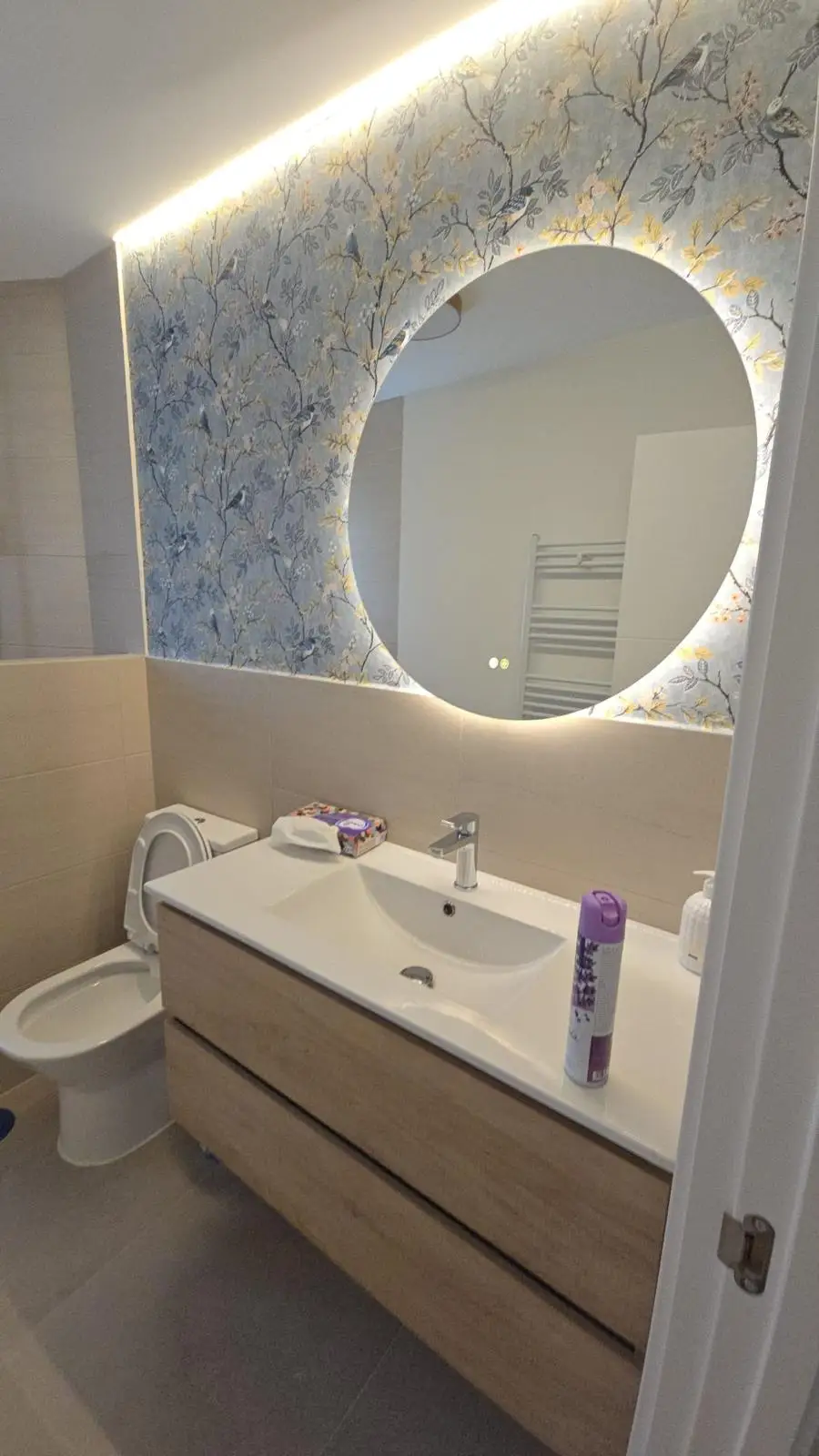 Reforma de baño con espejo circular retroiluminado sobre pared de papel pintado floral en Madrid