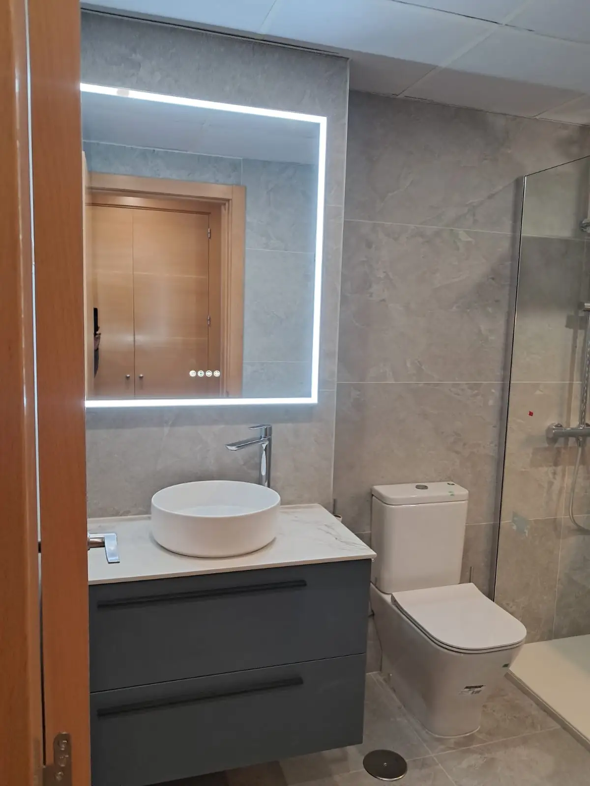 Reforma baño con mueble suspendido gris, lavabo sobre encimera y espejo retroiluminado en Madrid