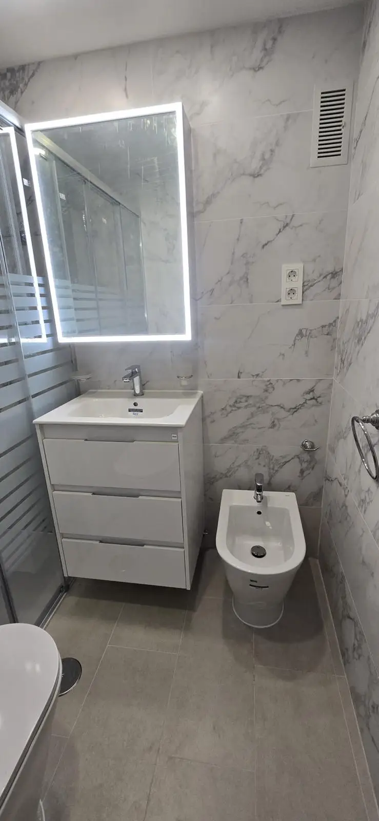 Reforma baño con revestimiento de mármol blanco, mueble suspendido y espejo retroiluminado en Madrid